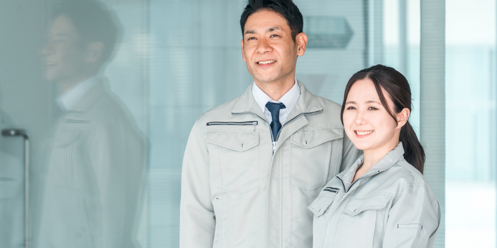 作業服を着た男性と女性社員がガラス壁の前で笑顔を向ける、プラスチック成形工場のスタッフ紹介写真