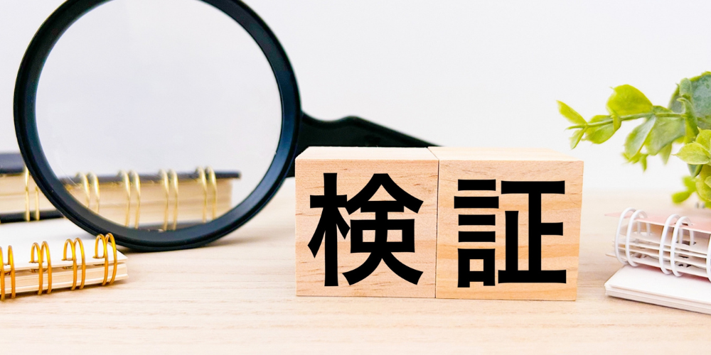 拡大鏡と木製ブロックに書かれた「検証」の文字、ノートと植物のあるデスク