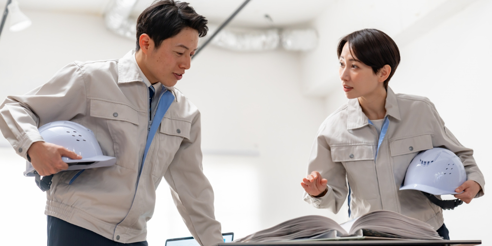 作業服を着た男女がヘルメットを持って図面を確認している様子