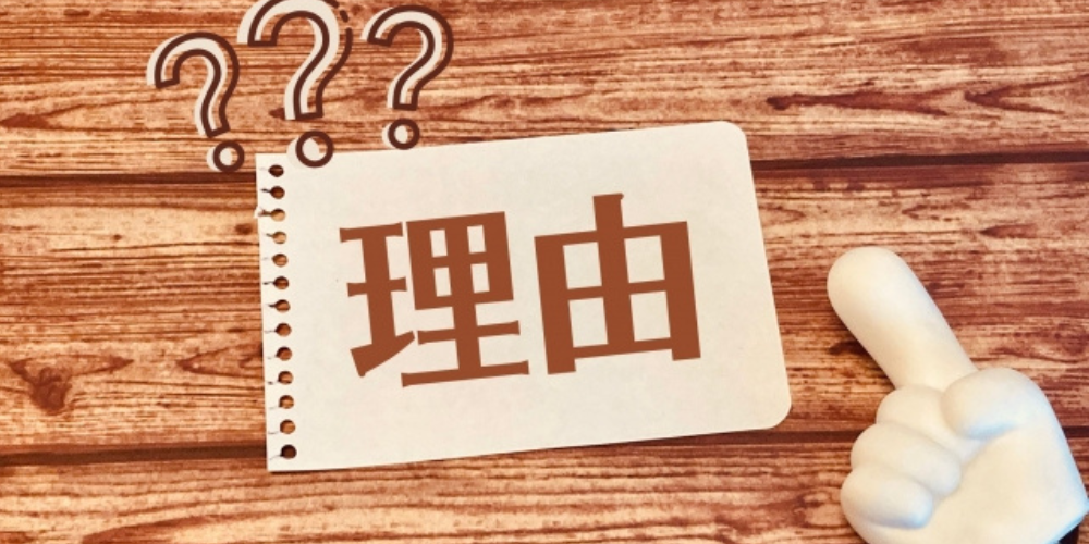木目の背景に「理由」と書かれたメモと疑問符、指差しポーズの手のオブジェクト