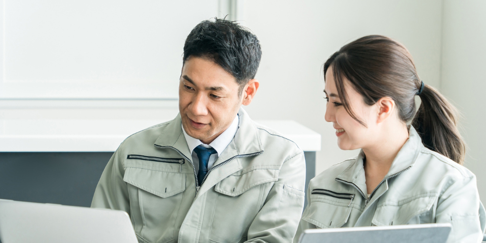 作業服姿の男女がパソコンとタブレットで会話する様子