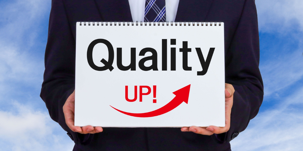 スーツ姿の人物が「Quality UP!」と書かれたボードを掲げているイメージ