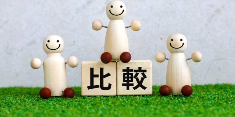 芝生の上に「比較」と書かれた木のブロックと、笑顔の木製人形が3体並んだ可愛らしい構成