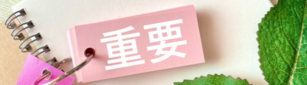 ピンクの単語帳に「重要」の文字があり、周囲にノートや葉っぱが並ぶ優しいトーンの構成