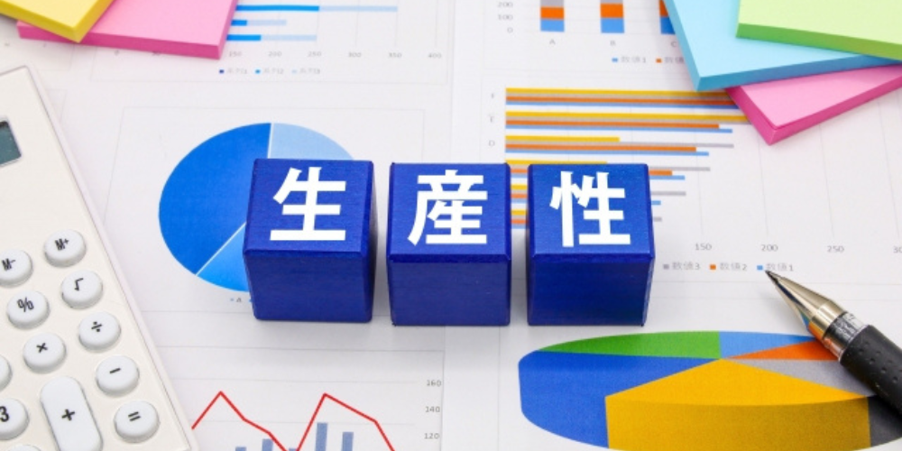 グラフや資料の上に「生産性」と書かれた青いブロック、電卓や文具が並ぶビジネスイメージ